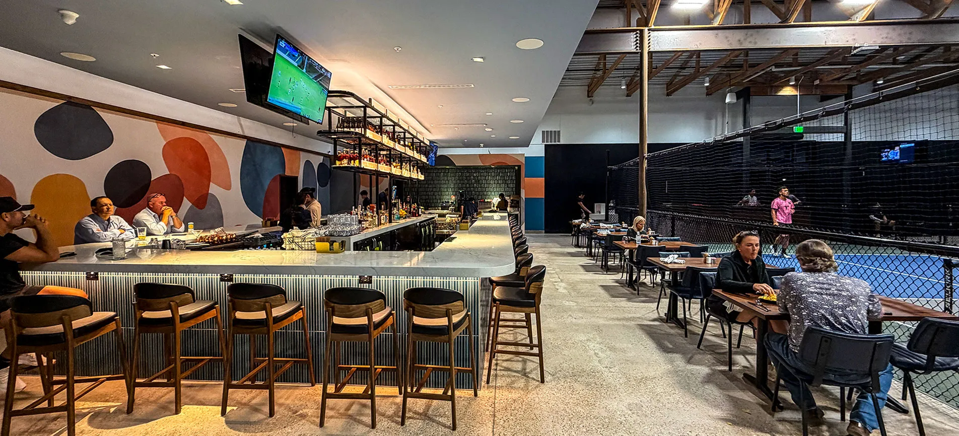 Inside Cork & Pickle: guests on stools at the long bar on the left, and tables along the netted pickleball courts on the right
