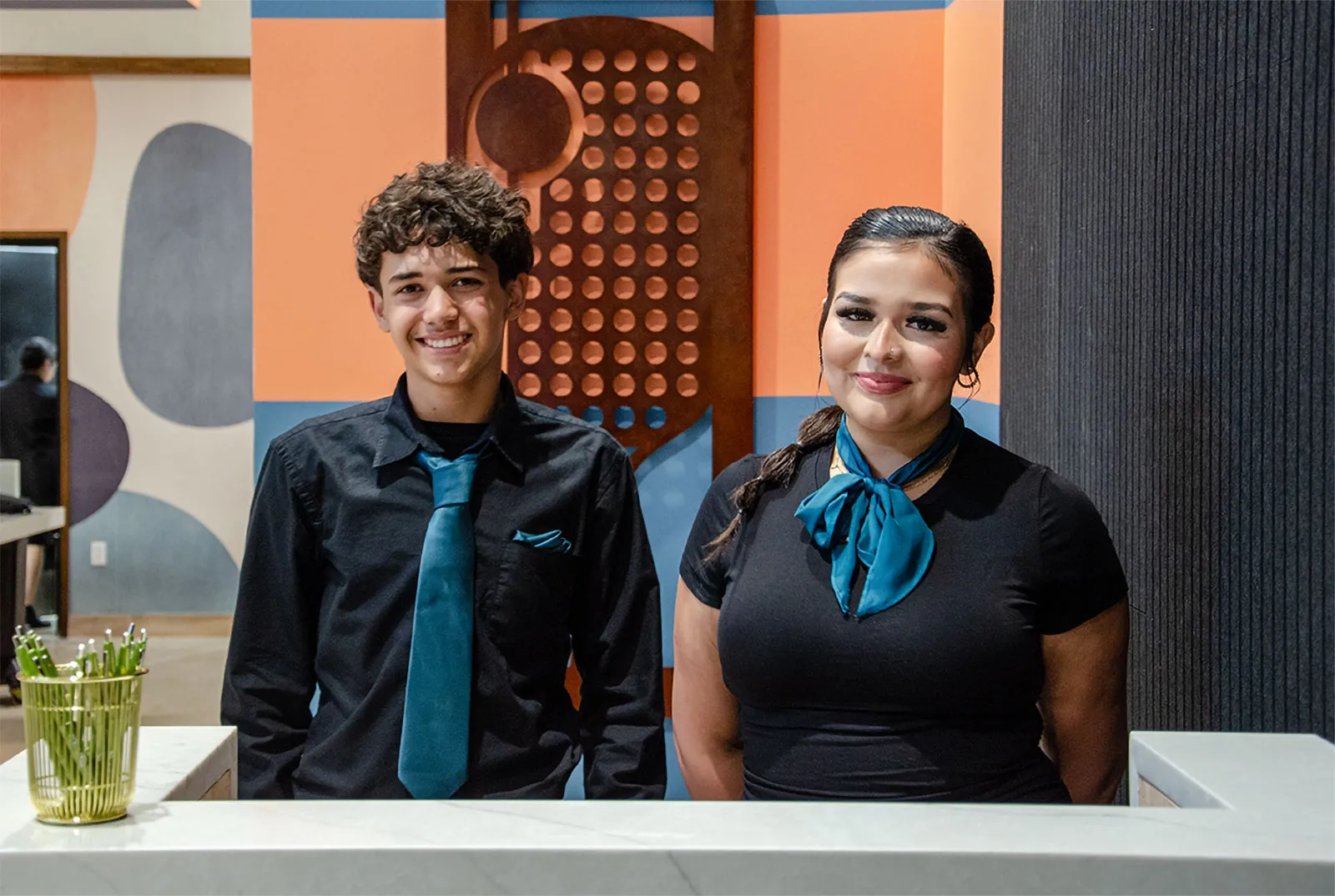 Two smiling Cork & Pickle hosts in black with teal ties, standing at the host stand