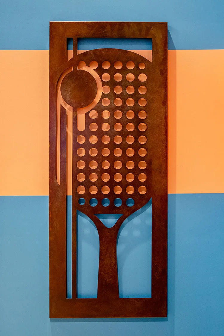 Rusted steel wall art of a pickleball paddle and ball on an orange and blue striped wall