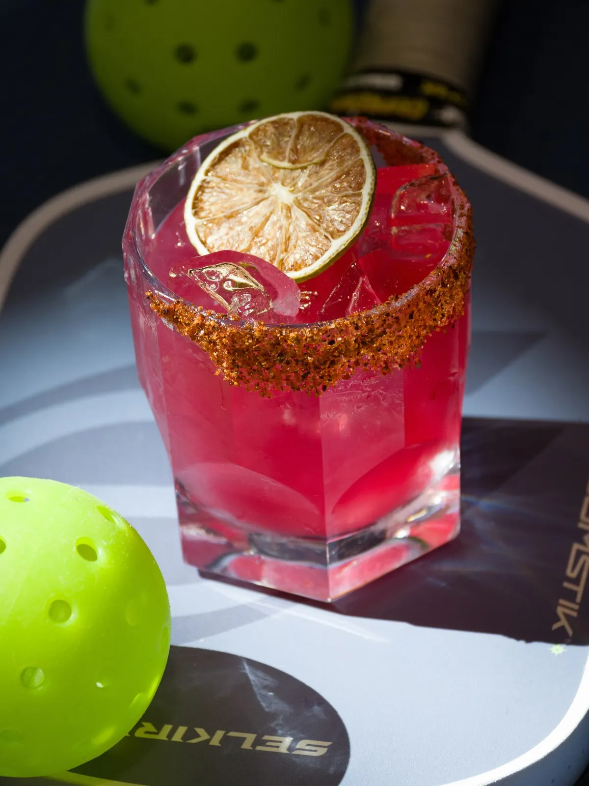 Pink cocktail with a chili-salt rim and a dried lime wheel, set on a pickleball paddle