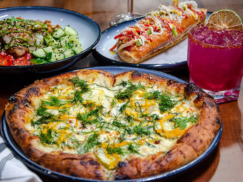 A Parmesan Pickle Pizza in front of a Power Bowl, a Street Dog, and a pink cocktail with a chili-salt rim