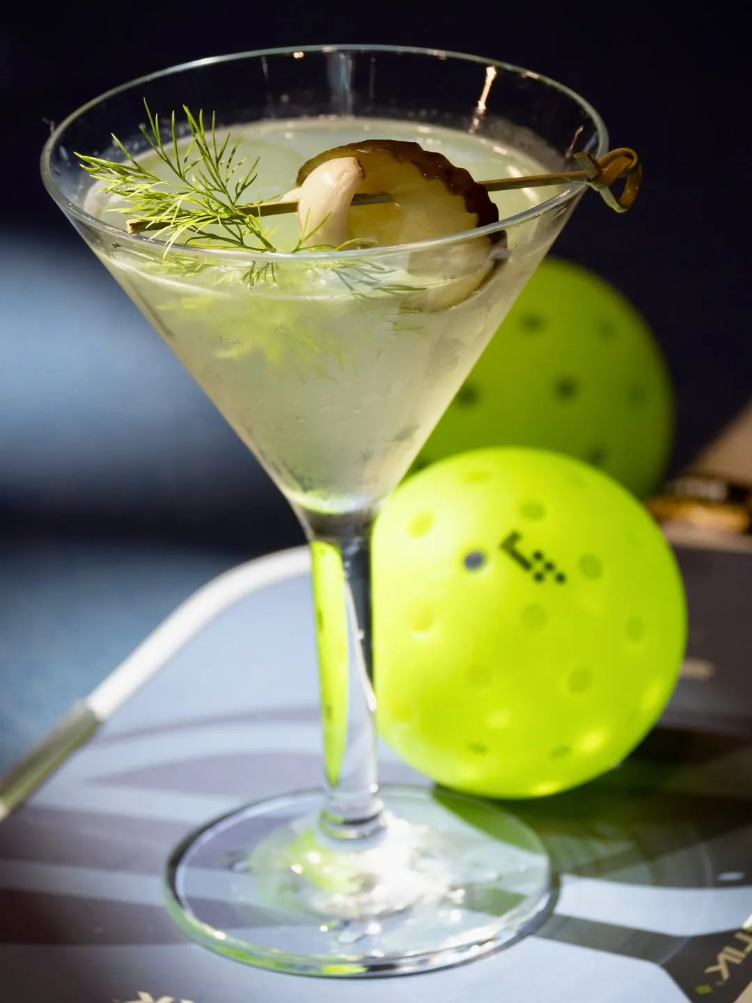 The Real Dill martini garnished with a pickle and a sprig of dill, beside a pickleball